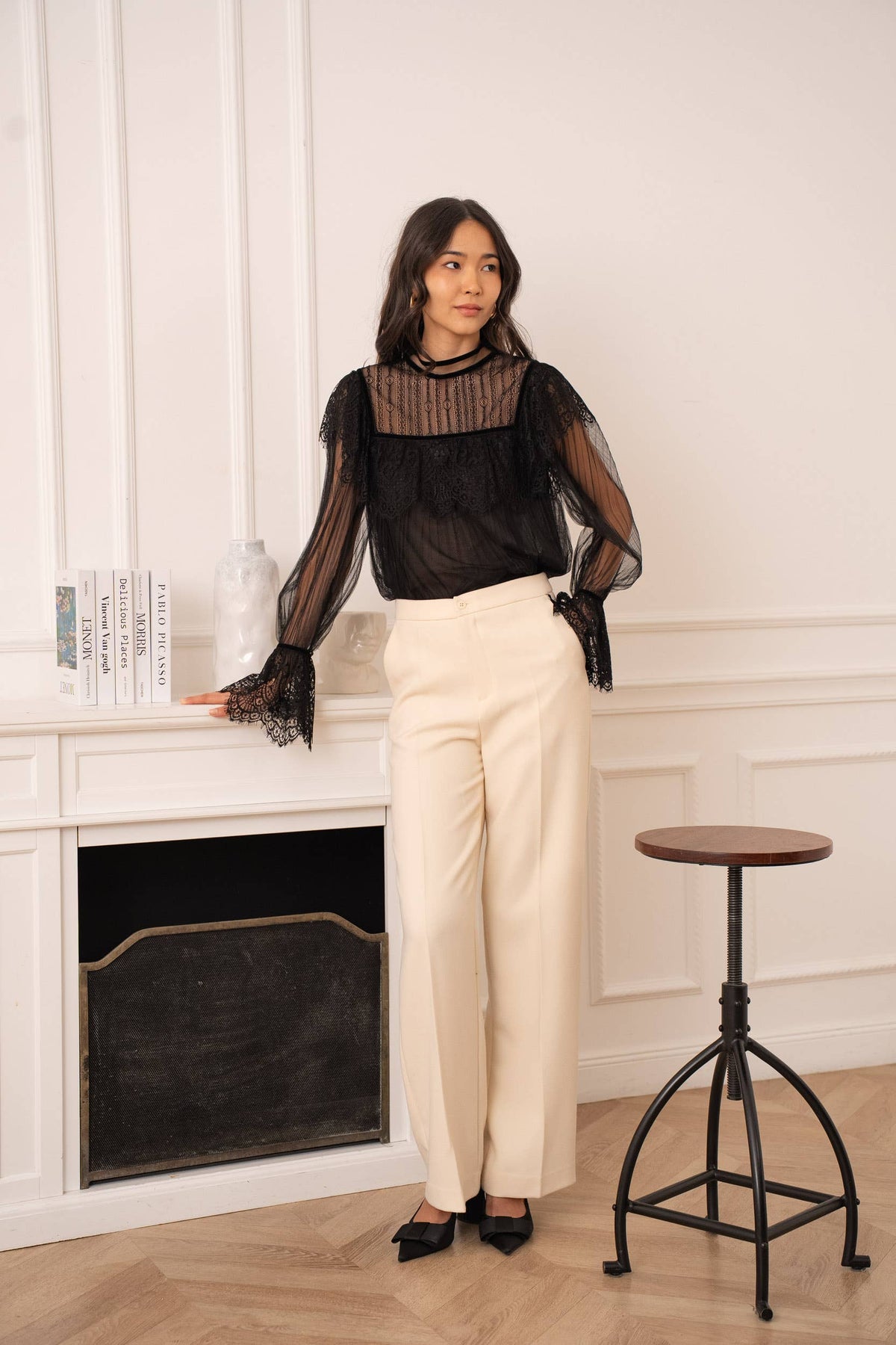 Woman wearing a black sheer blouse and white pants standing in a room with a fireplace and stool.
