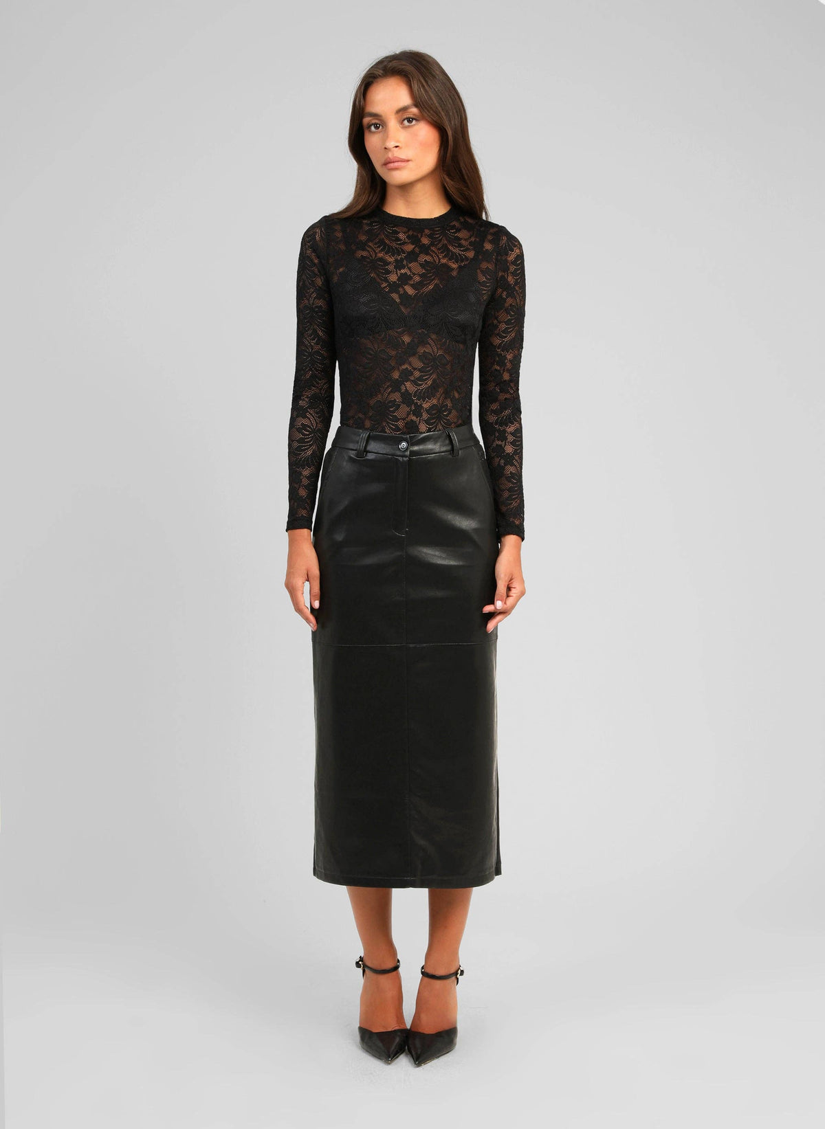 A woman modeling a black long-sleeve top with a sheer floral lace overlay and a black faux-leather midi skirt, paired with black pointed-toe heels, against a light gray studio background.
