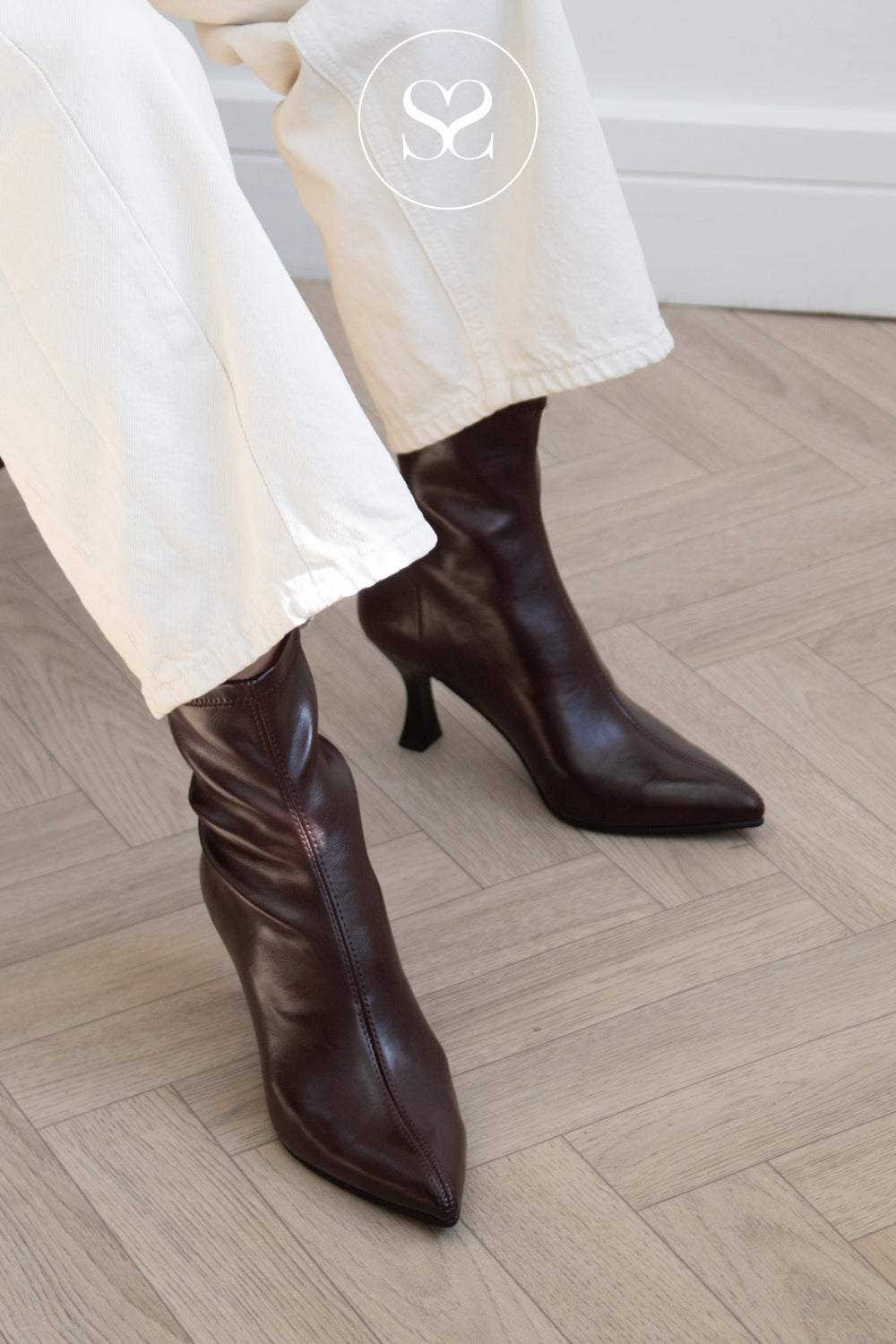A person wearing dark brown pointed-toe ankle boots with kitten heels and white pants, standing on a light wood herringbone floor. A circular logo watermark is visible near the top.