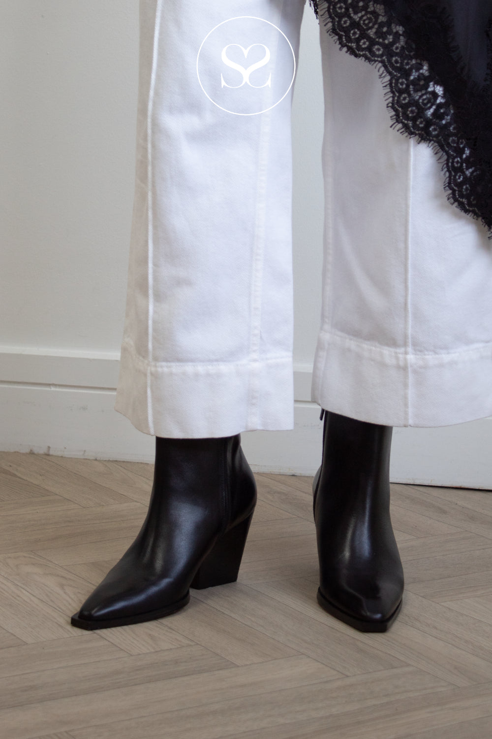 Angel Alarcon sleek Black leather pointed-toe ankle boots with a low block heel, shown on a model wearing white cropped trousers and a black lace-trimmed top, standing on a light wood herringbone floor.
