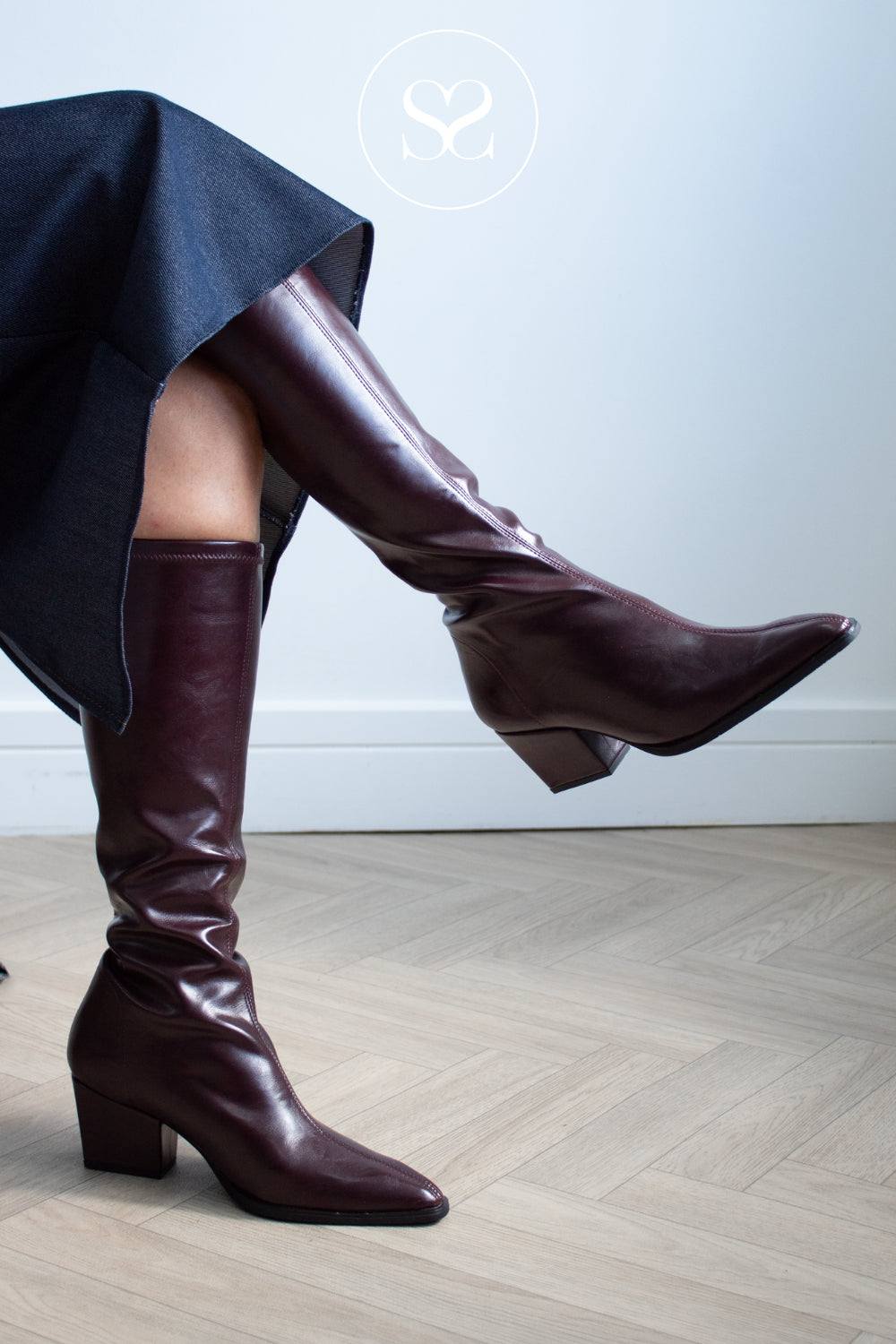 A pair of knee-high pointed-toe heeled boots in glossy deep burgundy, worn with a dark navy skirt. The miss elastic boots are shown on a model's legs against a light gray wall and light wood herringbone floor.