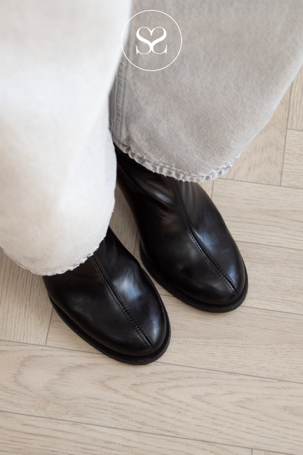 A close-up of a person wearing black leather ankle boots with light gray or off-white cropped pants, standing on a light wood floor. A circular 'SS' logo watermark is visible at the top center.