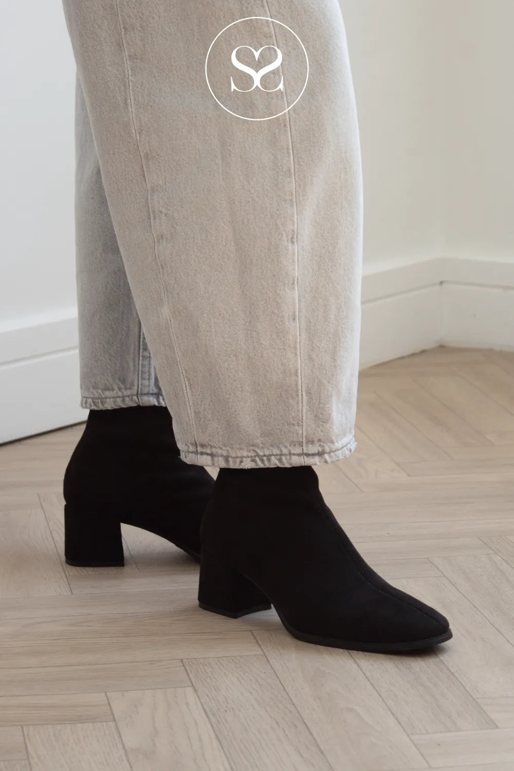 Black suede ankle height sockboots with block heels, shown on a model wearing light-wash jeans in a minimalist room with a light wood floor. Miss Elastic boots in Ireland