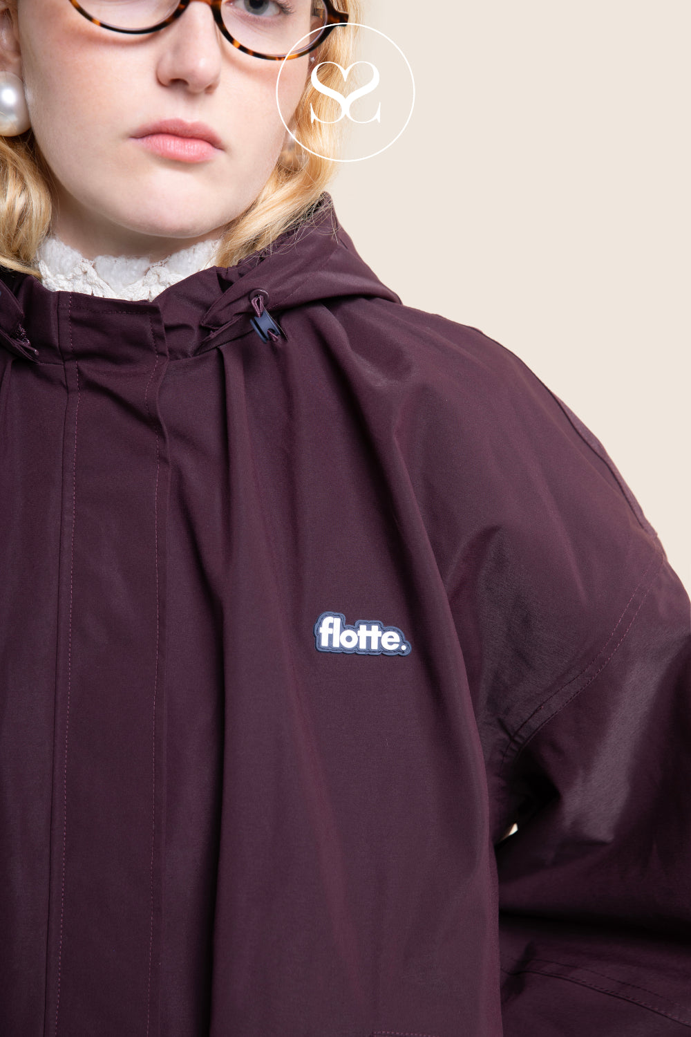 A close-up of a person wearing a dark plum hooded jacket with a small white and blue 'flotte.' logo on the chest, photographed against a plain light beige background.