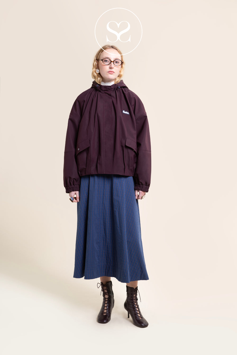 A model stands against a plain beige studio background wearing a dark Burgundy hooded jacket with front patch pockets, a long blue skirt, and dark lace-up ankle boots. A small white Flotte logo is visible on the left chest of the jacket.
