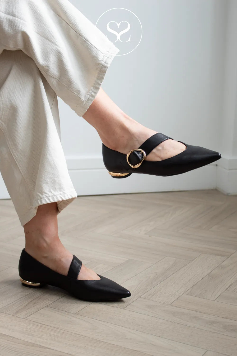 A person wearing black pointed-toe Mary Jane flats with gold-tone buckle and heel accents, paired with light beige cropped pants, standing on a light wood herringbone floor.