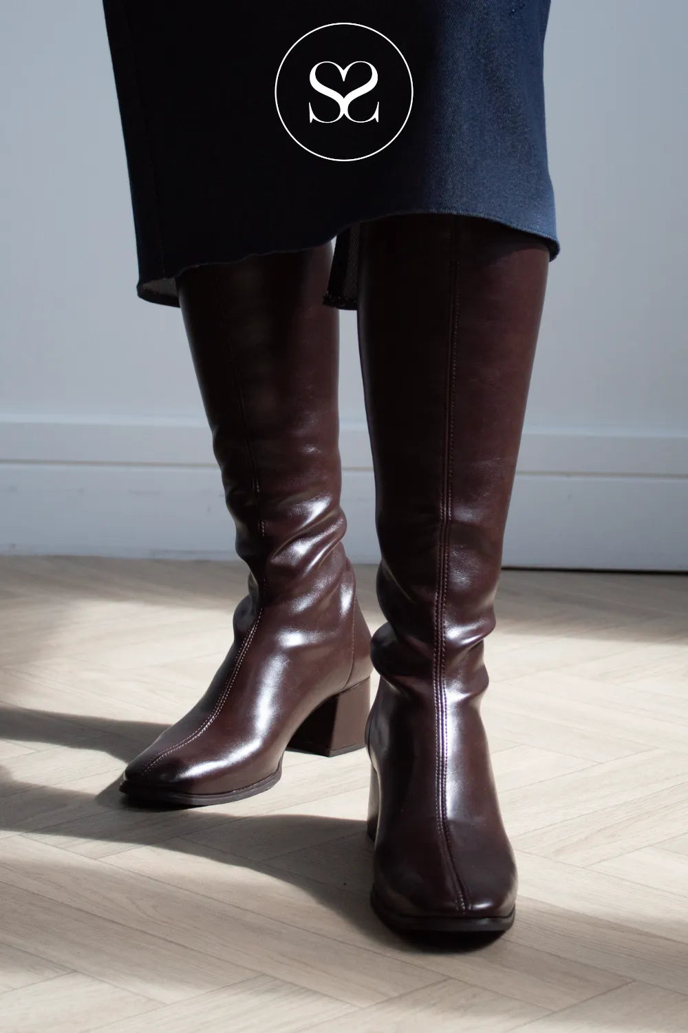 A person wearing dark chocolate brown knee-high leather boots with a block heel, shown from the knees down in a brightly lit room with light wood flooring and a white wall. A circular logo with an 'Sheneil Shoes' is overlaid near the top.