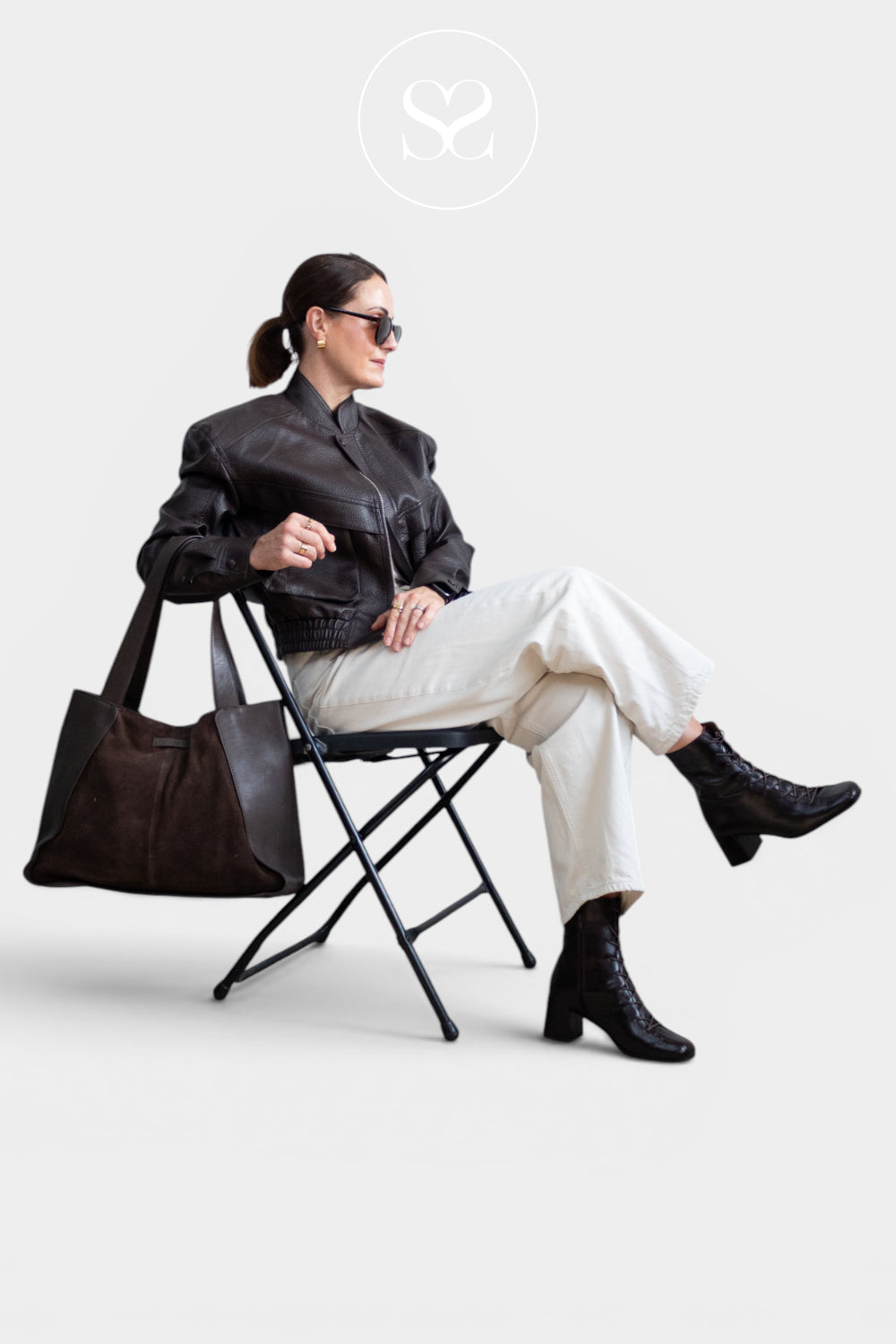 Editorial image: A woman sits on a black folding chair wearing a black leather jacket, off-white trousers, and black ankle boots, with a Depeche large dark brown tote bag resting on the chair. She is wearing dark sunglasses against a light grey studio background.