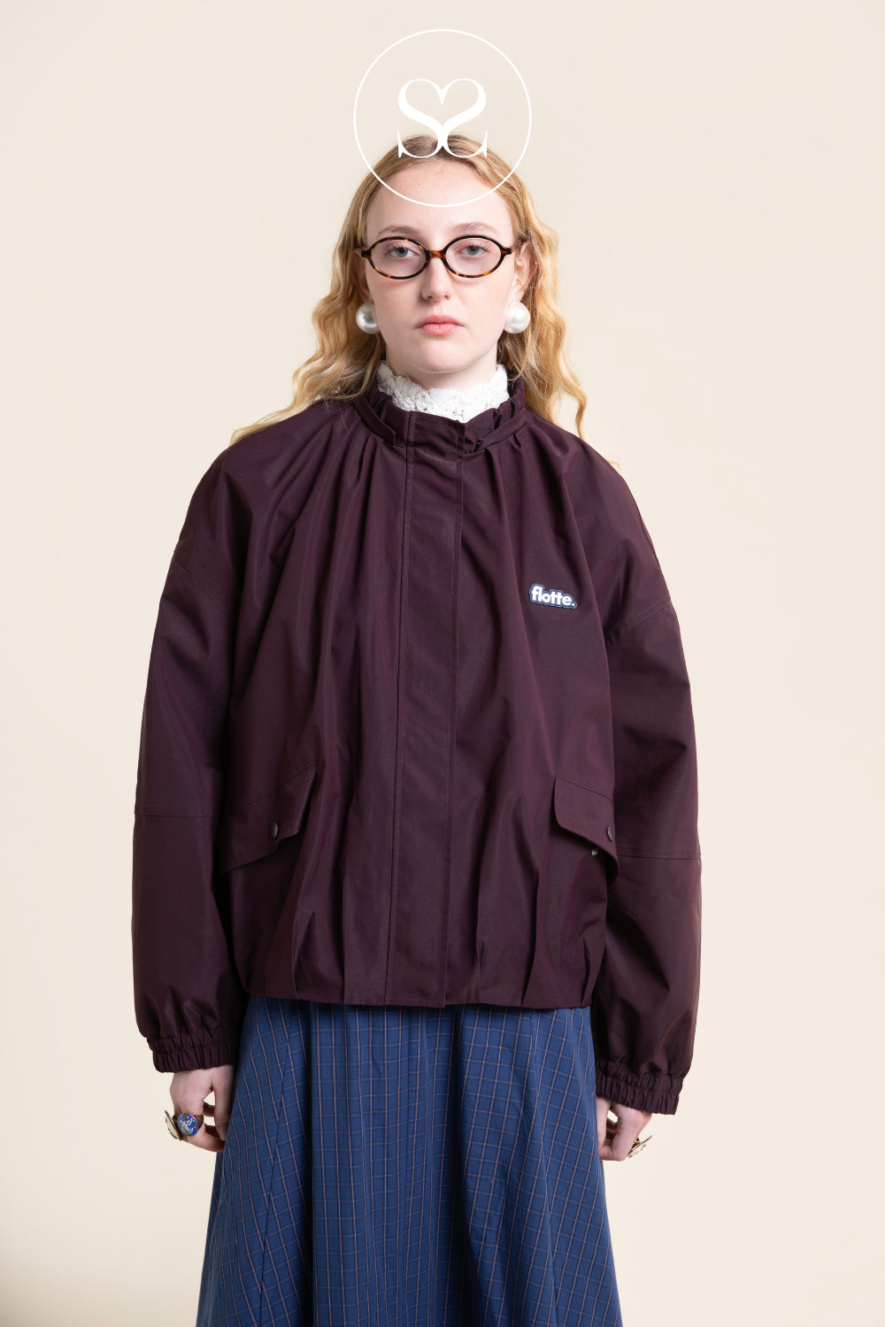A model wearing a dark BURGUNDY windbreaker jacket with a white embroidered Flotte logo on the chest, paired with a blue pinstripe skirt and glasses, set against a solid beige background.