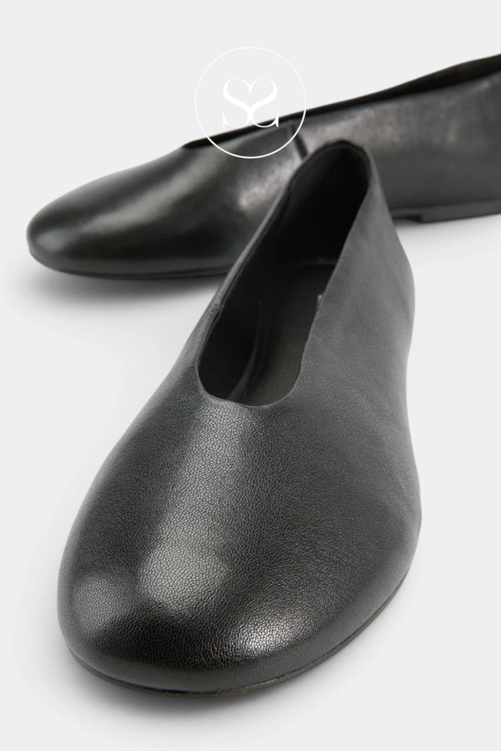 A pair of black leather ballet flats with a rounded toe, shown on a light gray background. One shoe is in sharp focus in the foreground, while the other is softly blurred in the background.