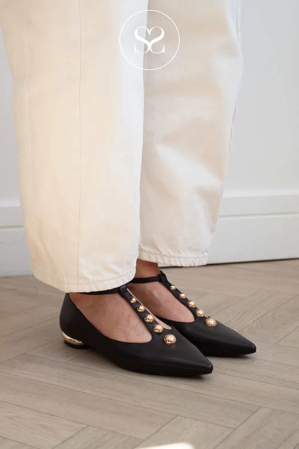 Black pointed-toe Mary Jane flats with gold-tone decorative studs and a low block heel, shown on a model wearing cream trousers on a light wood floor.