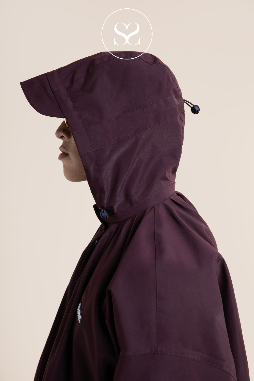 A side-profile view of a person wearing a Flotte hooded raincoat against a light beige studio background. The coat has a smooth matte finish, a high hood with a drawstring, and a drawcord hem.
