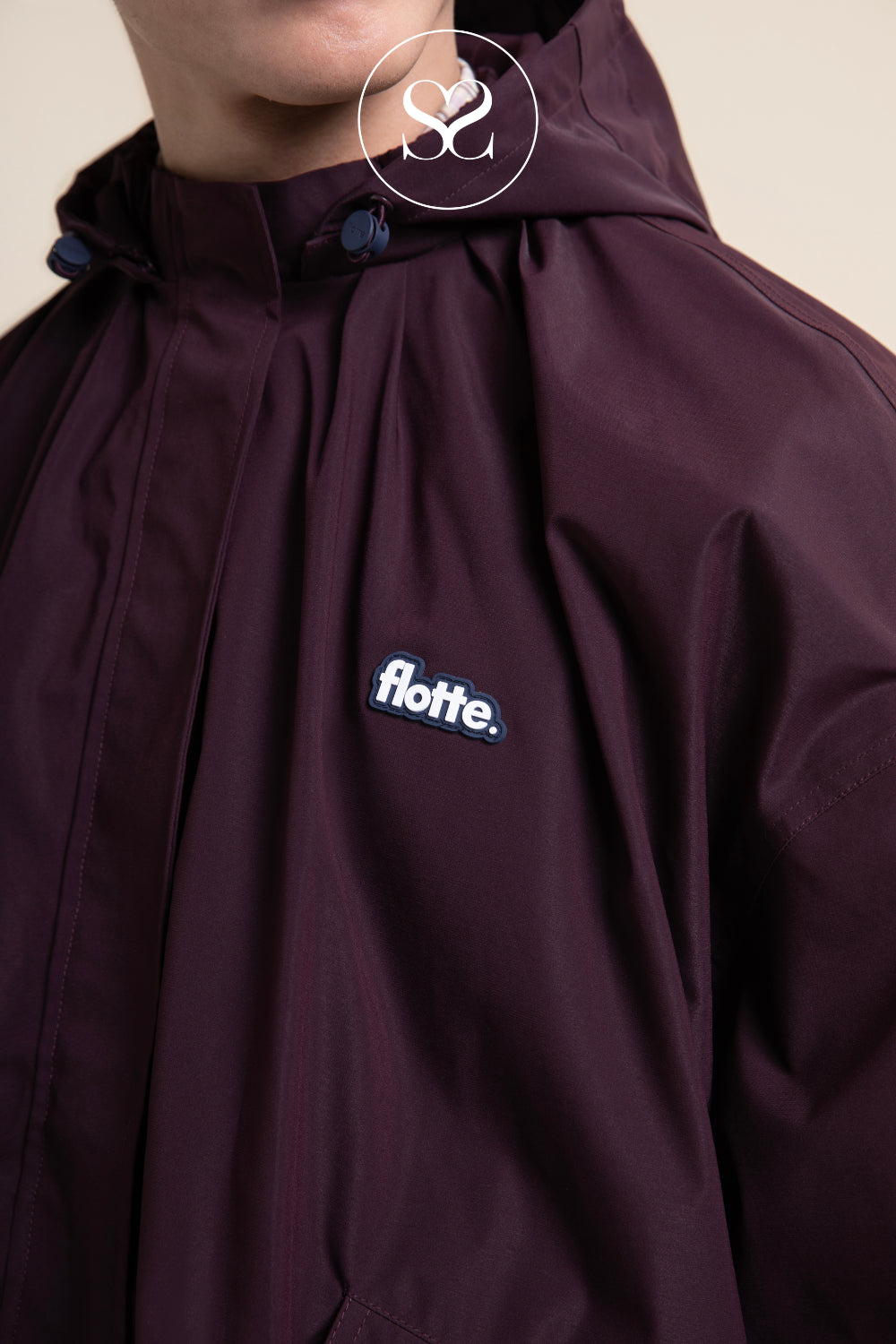 Close-up of a person wearing a dark plum hooded jacket with a visible 'flotte.' chest logo, photographed against a plain light beige background.