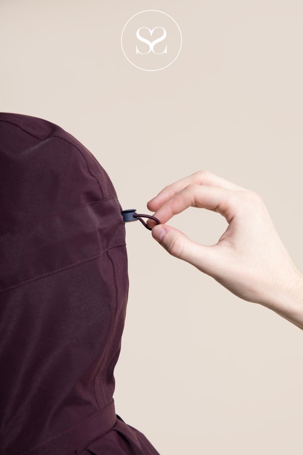 A close-up of a person's hand adjusting the drawstring of a dark plum hooded garment against a solid beige background.