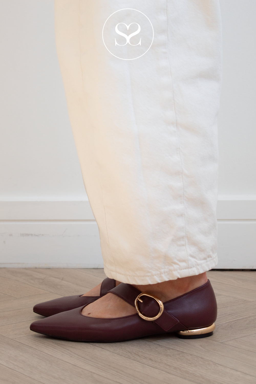 A person wearing off-white trousers and Gioseppo burgundy pointed-toe Mary Jane flats with gold-tone buckle straps, standing on a light wood floor against a white wall. Photographed by sheneil shoes Galway Ireland