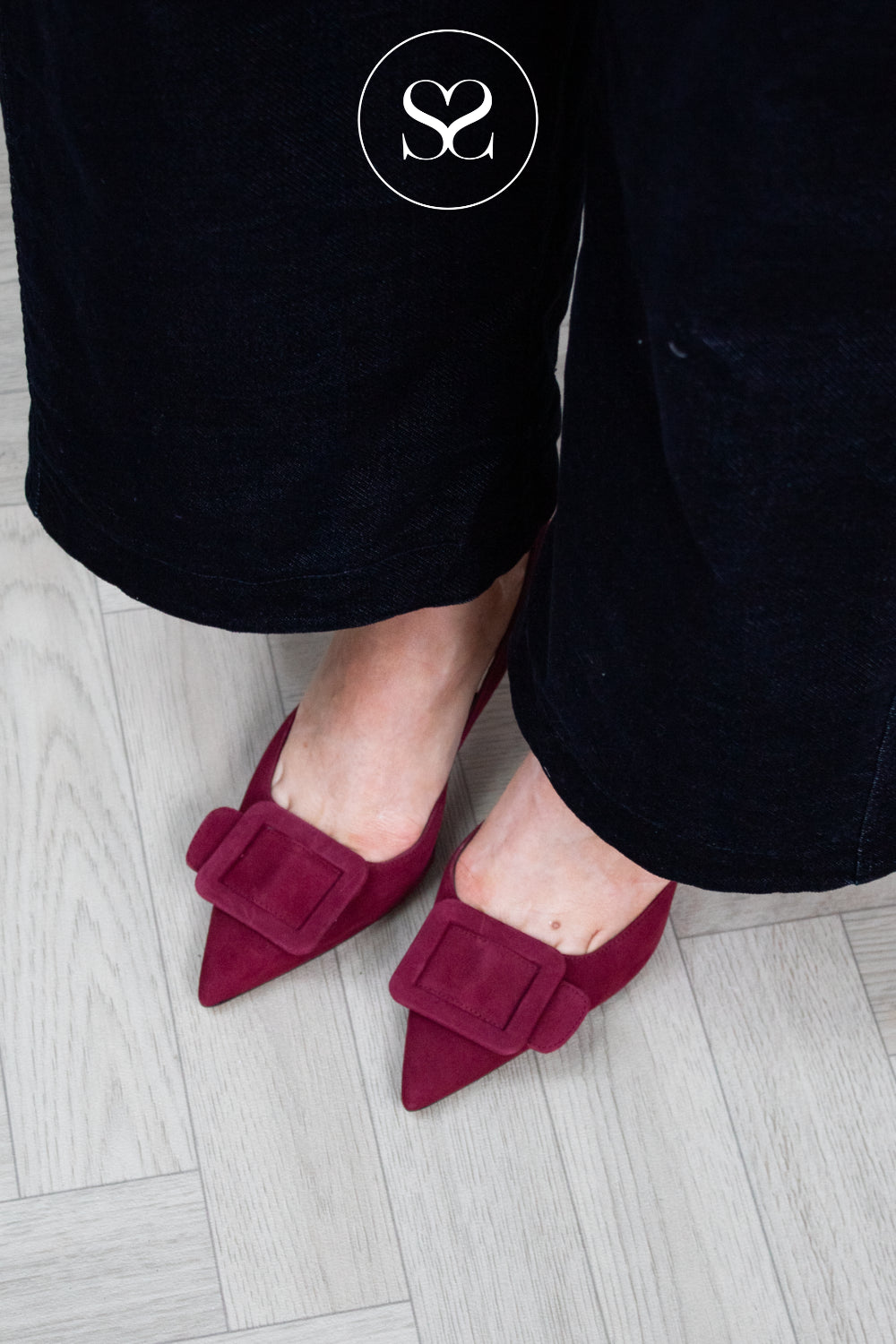 A close-up of a person wearing dark burgundy pointed-toe heels with large square buckle details, paired with navy velvet wide-leg pants on a light gray wood floor. A white circular logo with an 'sheneil shoes' is visible near the top. Shoes are lodi nestor