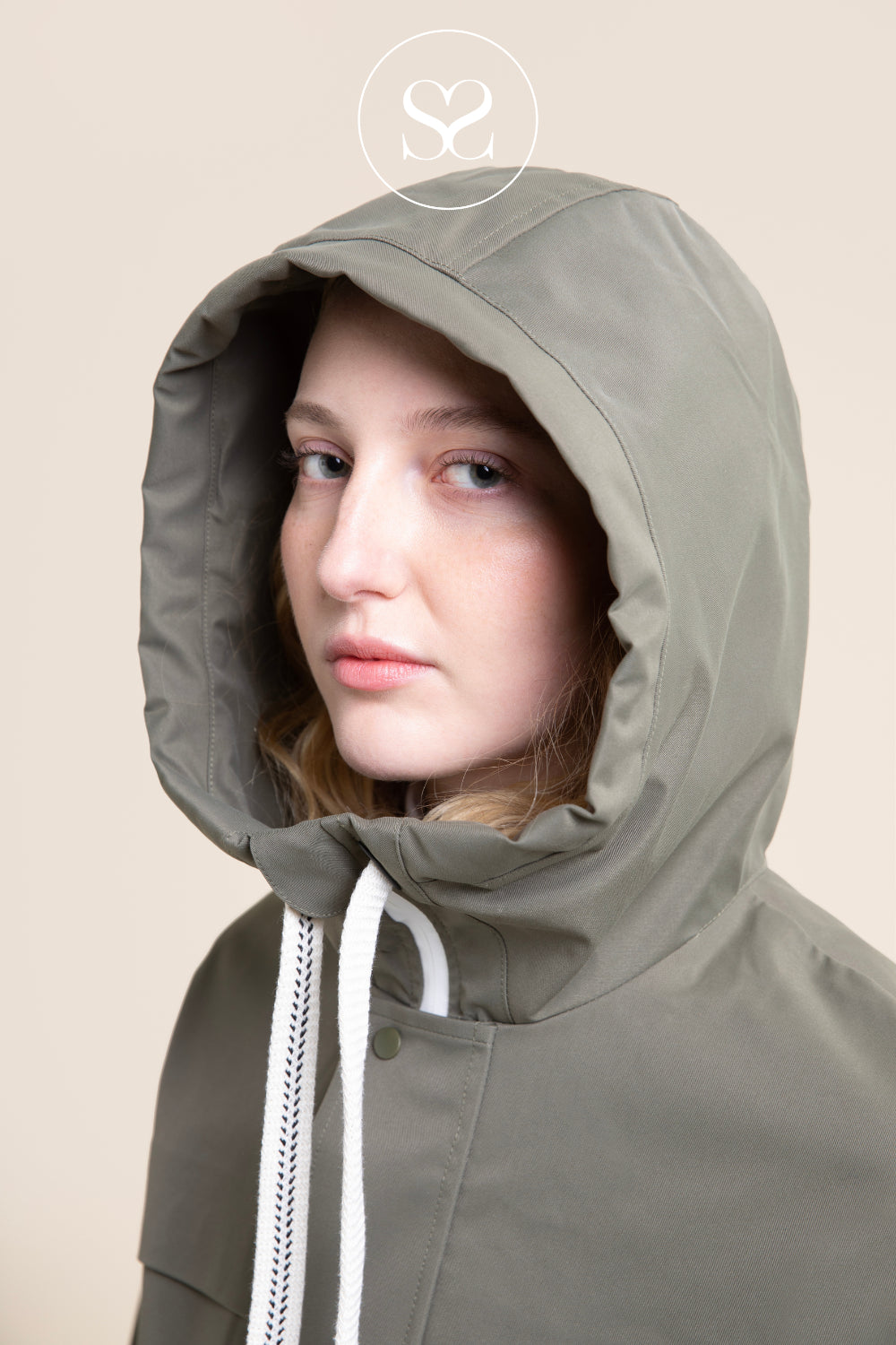 A close-up of a model wearing a khaki hooded jacket with white drawstrings, set against a solid beige background.