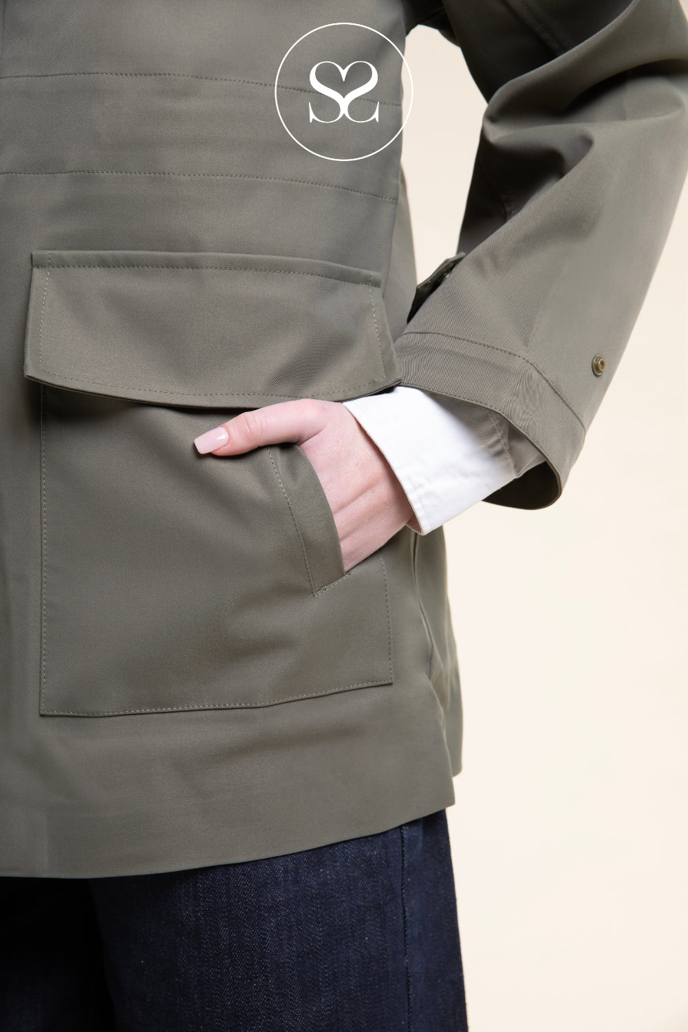 Close-up of a person wearing an olive green utility jacket with a large front flap pocket, a white shirt cuff visible at the pocket, and dark blue jeans against a light beige background.
