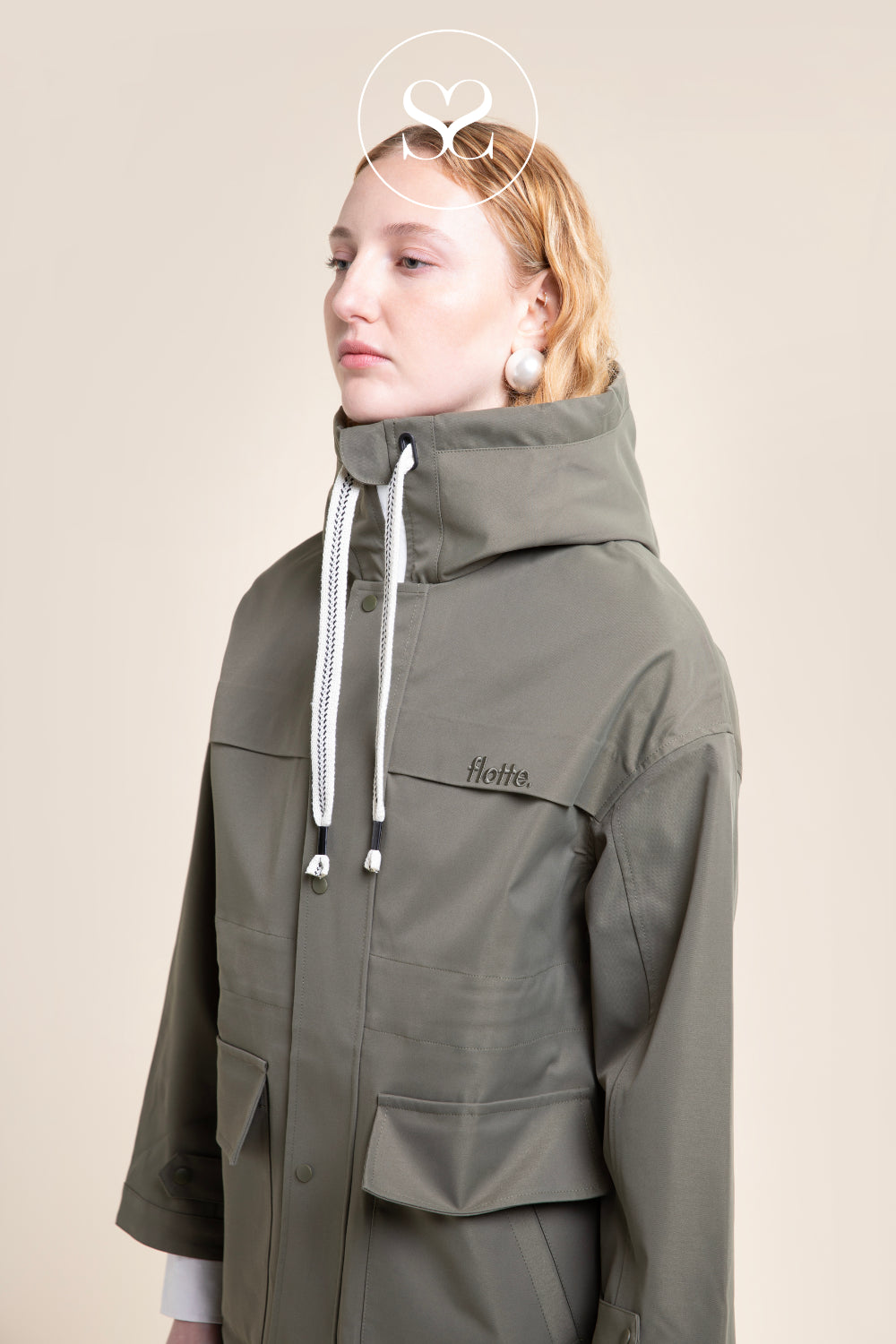 A woman wearing an olive green hooded jacket with white drawstrings and a small 'Flotte' brand logo on the chest, photographed against a plain beige studio background.