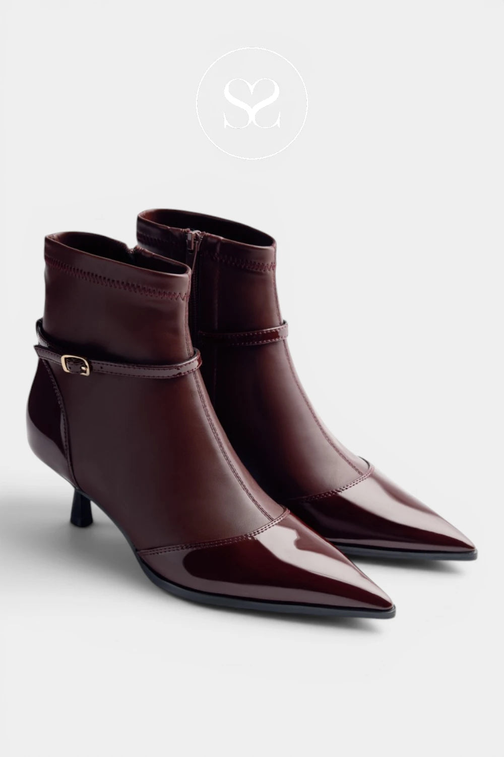 A pair of chocolate brown pointed-toe ankle boots with a glossy patent leather finish, a side buckle strap detail, and a kitten heel, shown on a light gray background