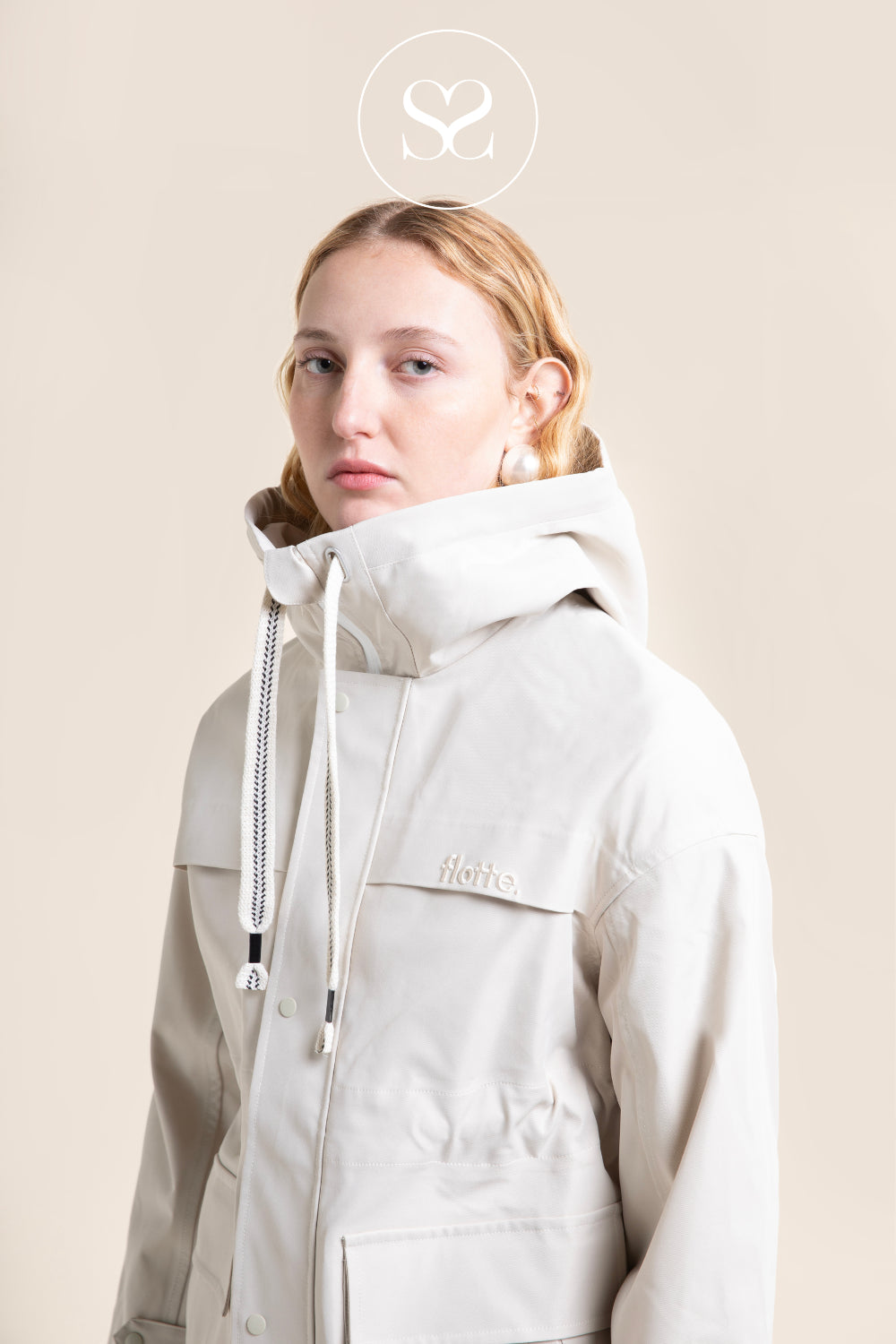 A model wearing an ivory hooded waterproof jacket with a high collar, front zipper, and drawstrings, set against a solid beige background. A circular logo with an 'S' is visible at the top of the image.