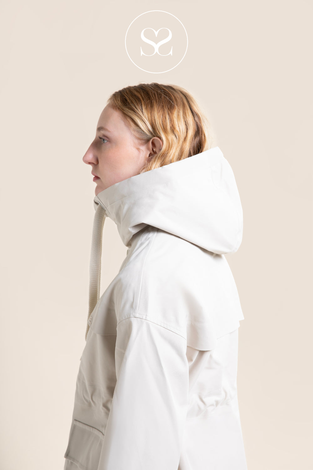 A woman in profile wearing a light ivory hooded funnel neck jacket, shown against a solid light beige background with a circular sheneil shoes logo overlay at the top.