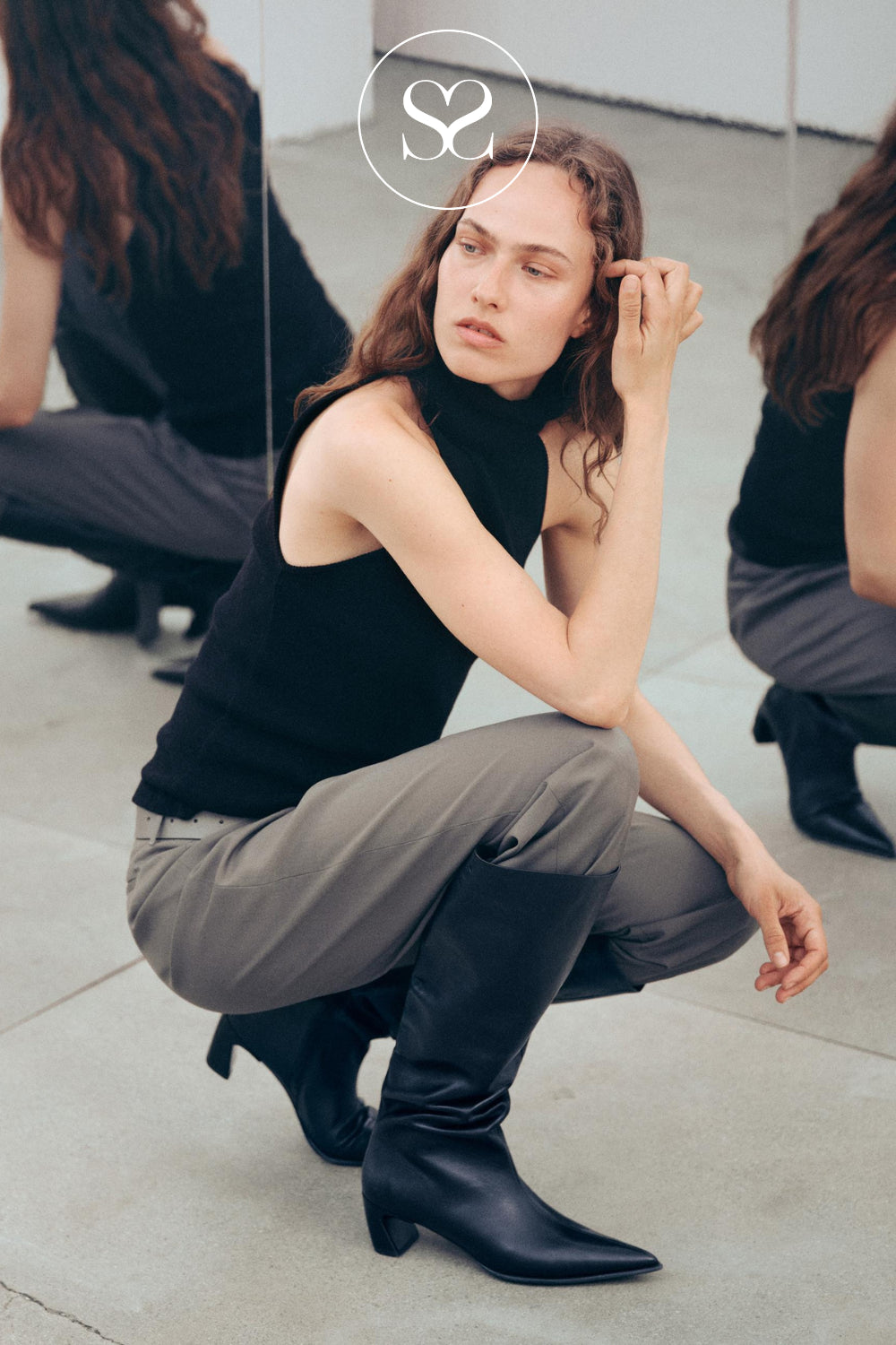 Black pointed toe knee high boots with curved heel and slouch fit worn with grey trouser in studio - Vagabond shoemakers style