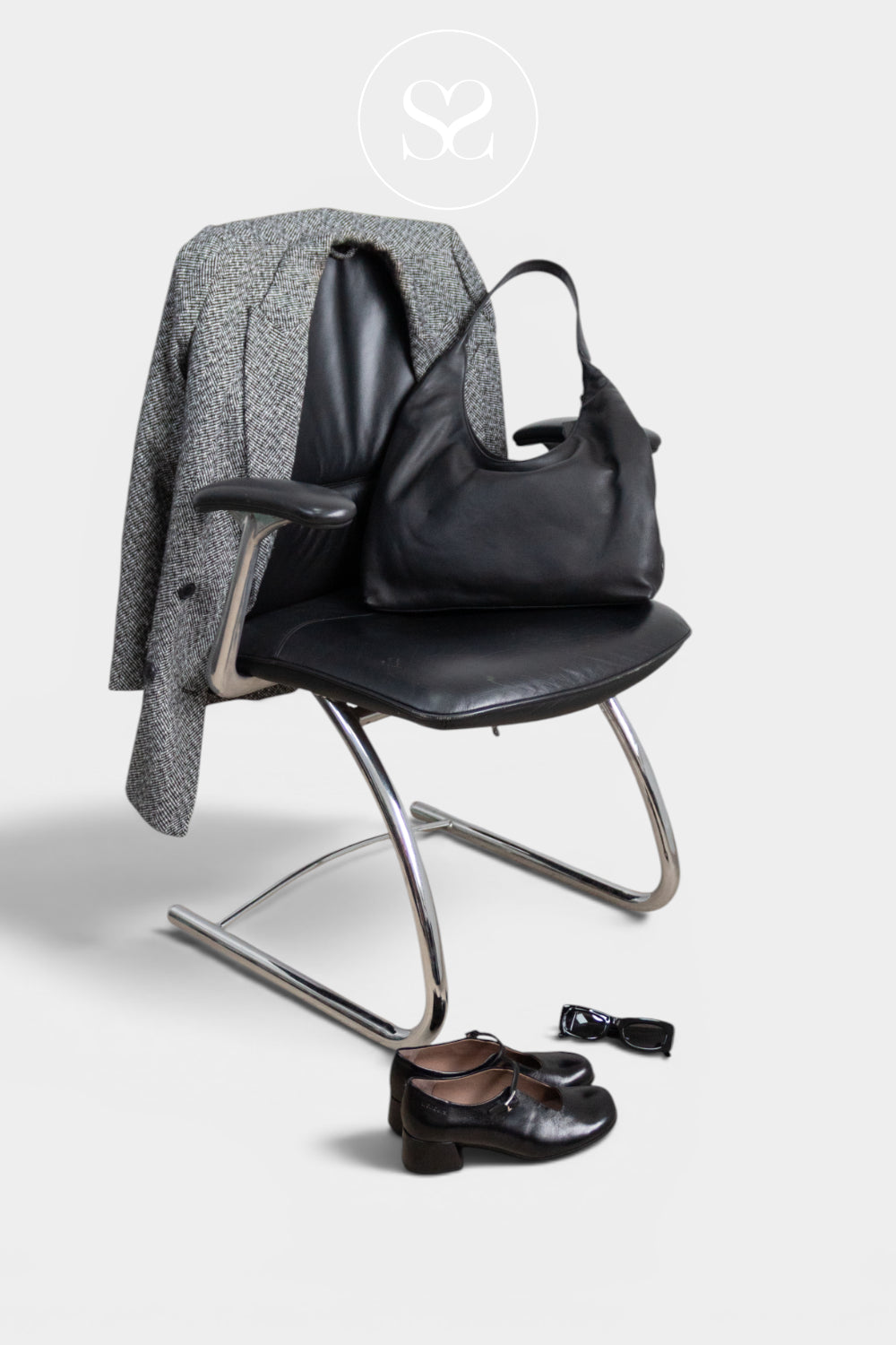 A depeche black leather hobo handbag sits on a black office chair next to a gray patterned blazer draped over the chair arm, with a pair of black leather loafers placed on the floor in front. The scene is photographed on a clean, light grey background with a faint circular Sheneil Shoes logo watermark at the top. Location Ireland