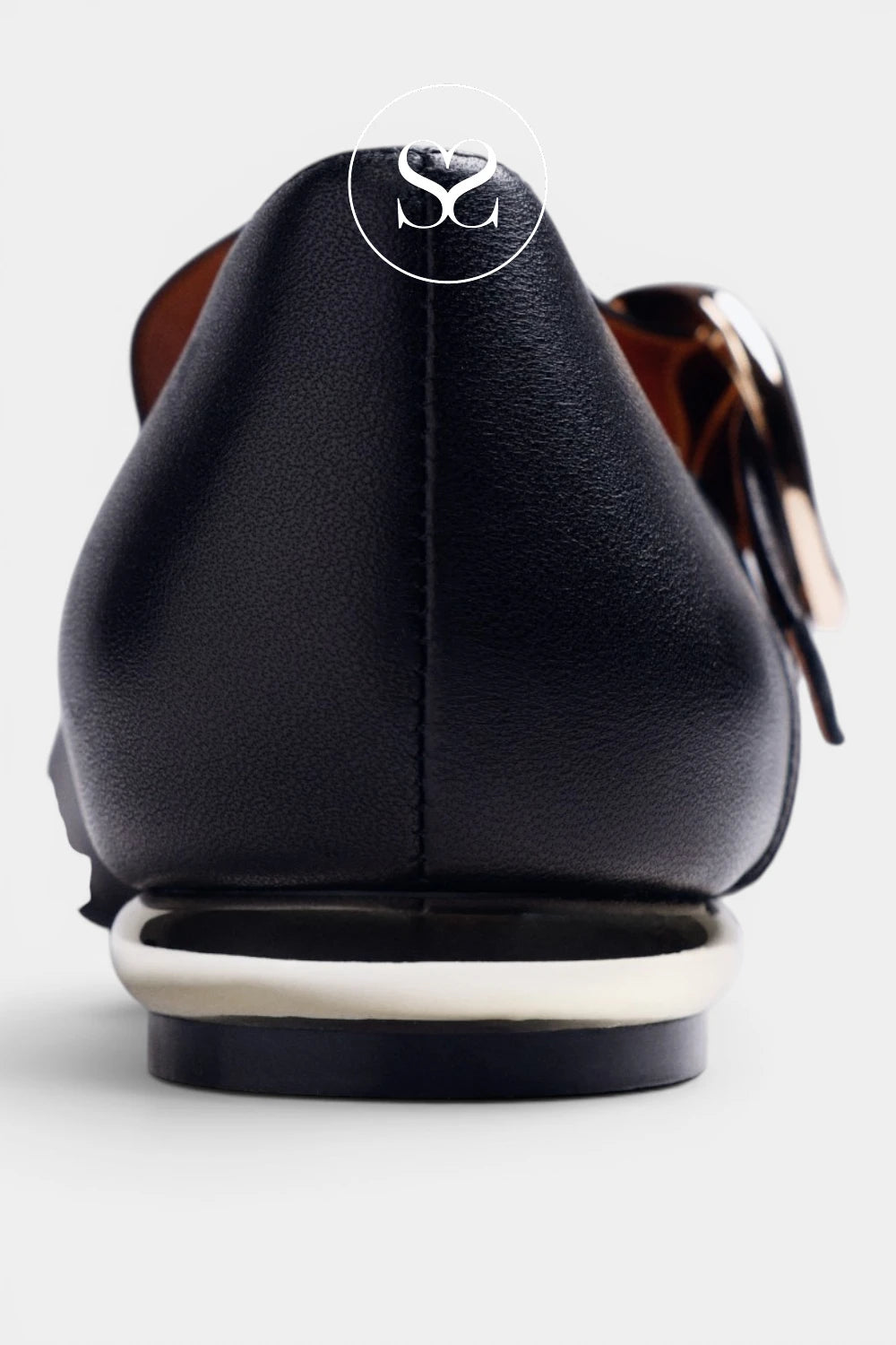 Close-up rear view of a black leather shoe with a chunky white sole and a metallic gold decorative accent on a light gray background.