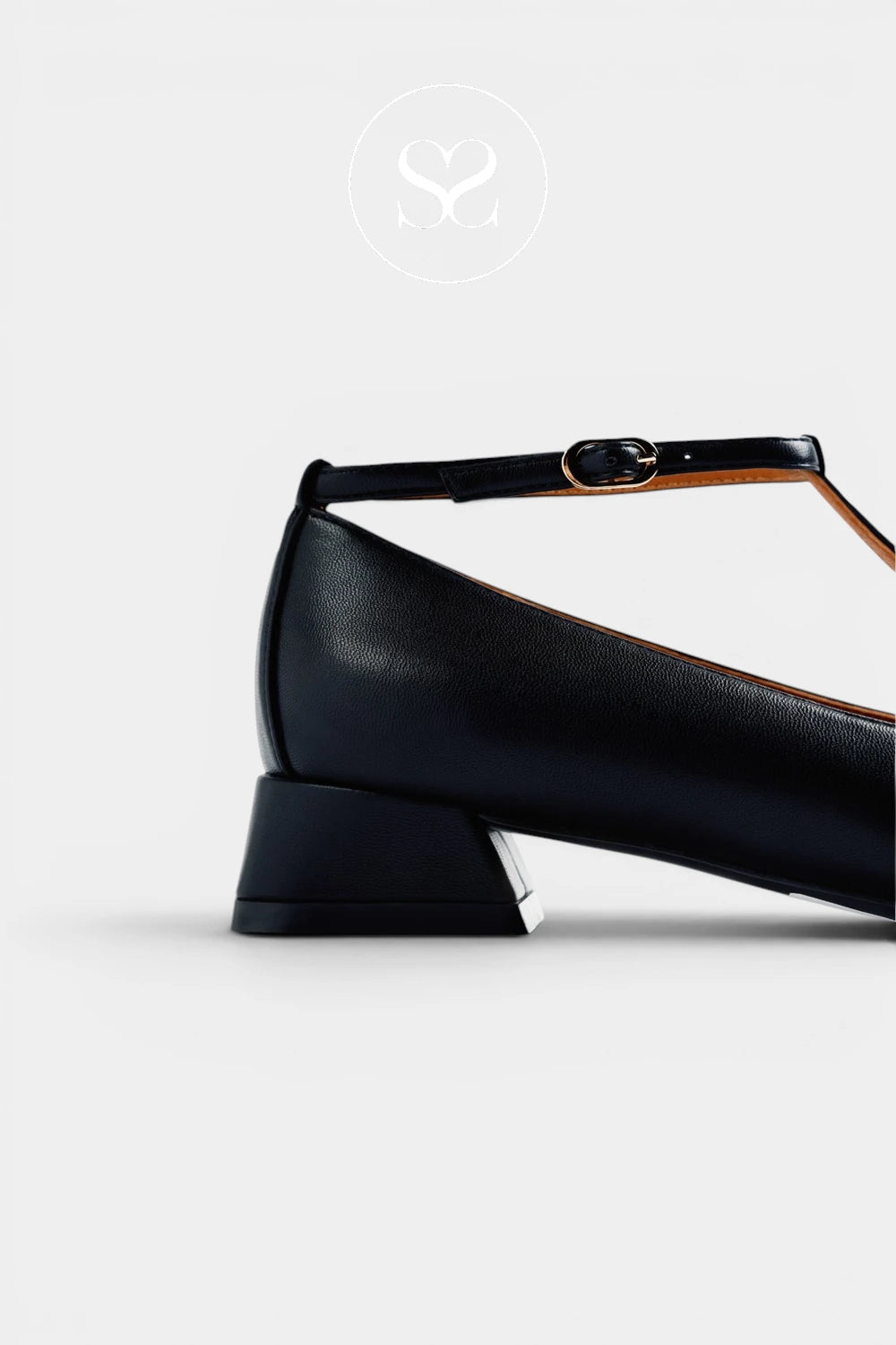 A close-up side view of a black leather T-strap shoe with a low block heel and a small gold-tone buckle, shown on a light gray background with a faint logo above.