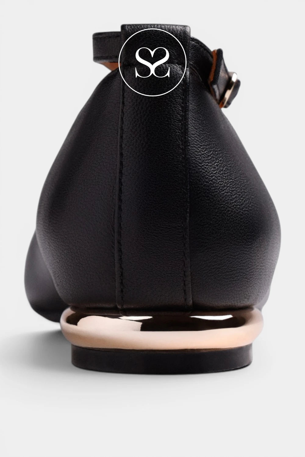 A close-up rear view of a black leather flat shoe with a metallic rose gold circular heel accent and a black ankle strap featuring a white circular logo with an 'S' monogram.