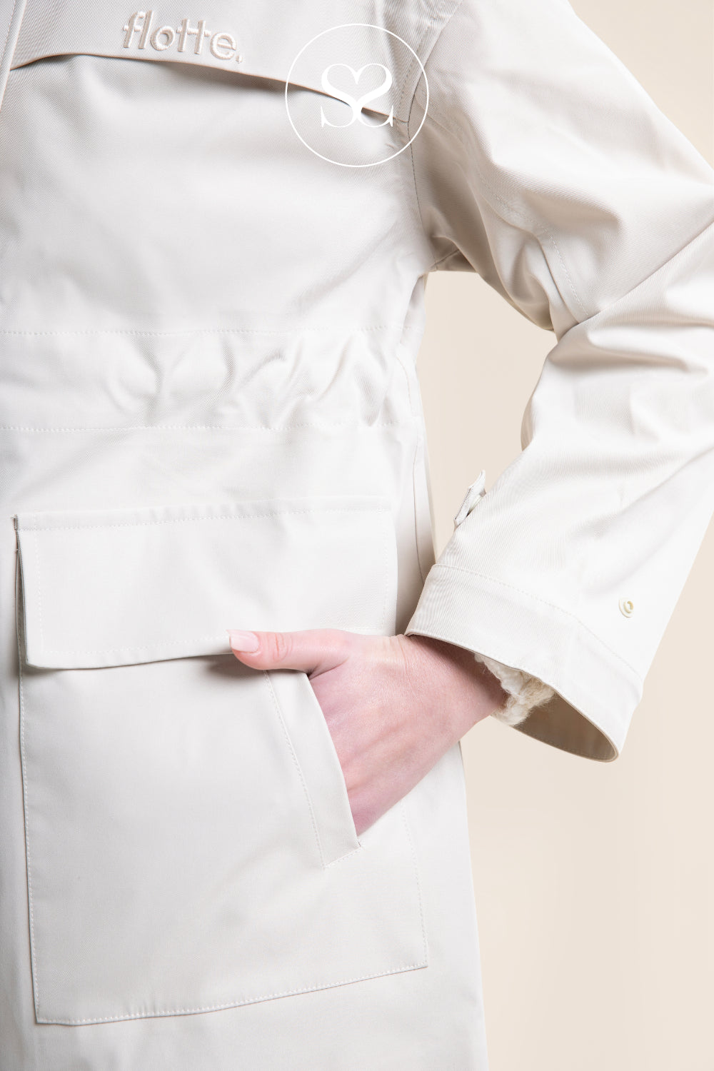 Close-up of a person wearing a light beige utility jacket with a flap pocket and a hand tucked into the pocket. The brand name 'flotte.