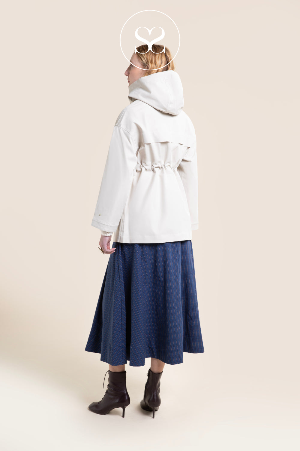 A woman shown from the back wearing a white hooded jacket with a drawstring waist from Flotte, a navy midi skirt, and dark heeled ankle boots, against a plain beige studio background in Ireland