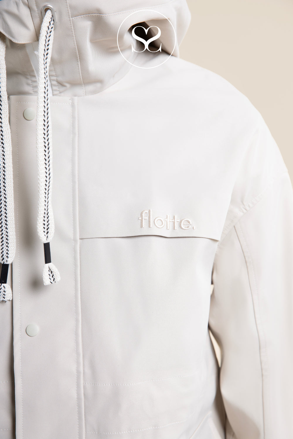 Close-up of a light cream hooded jacket with white drawstrings and a small tonal 'flotte.' logo on the chest, shown against a warm beige background.