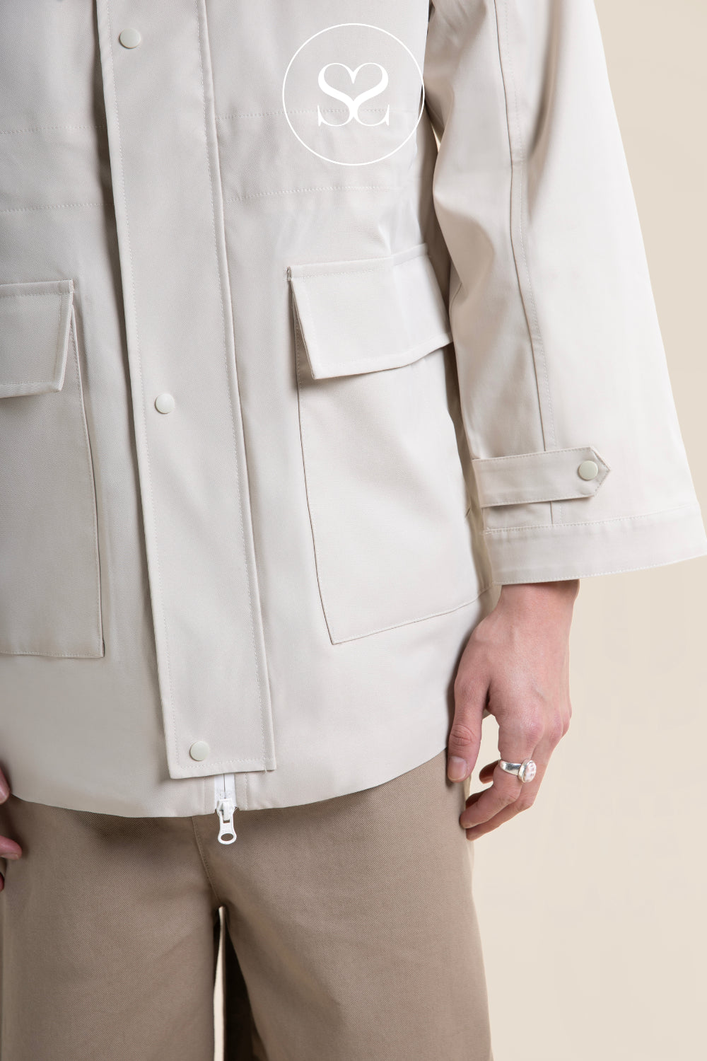 A close-up of a person wearing a light beige utility jacket with flap pockets and a small circular logo on the chest, paired with beige pants against a plain beige background.