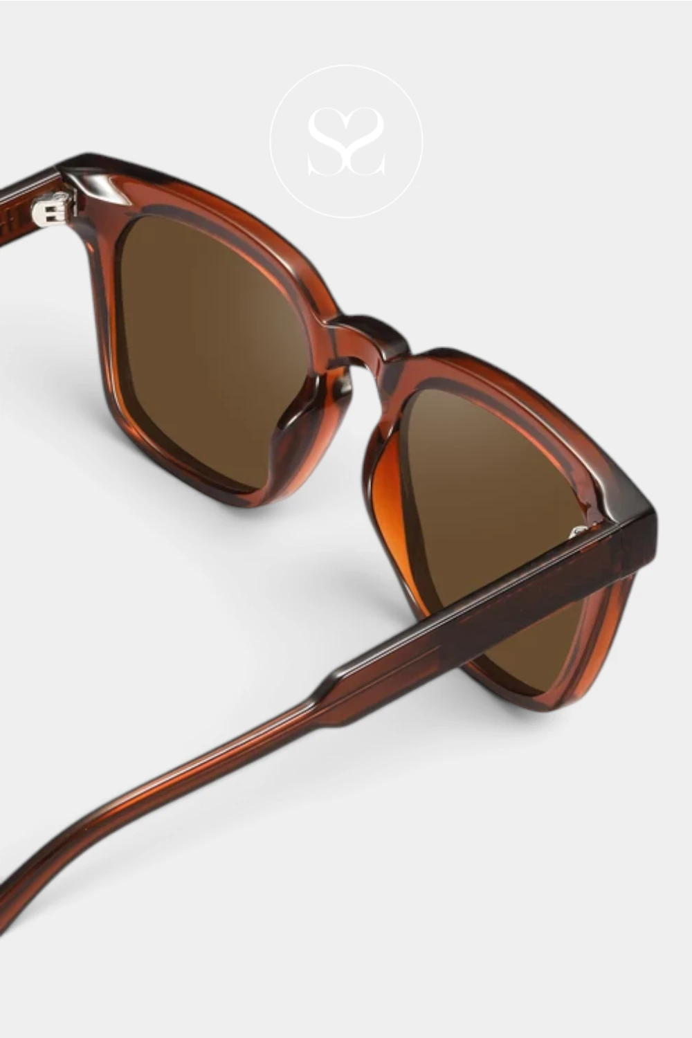 Close-up of a pair of sunglasses with a translucent amber-brown acetate frame and brown tinted lenses, shown on a light gray background with a faint circular logo above.