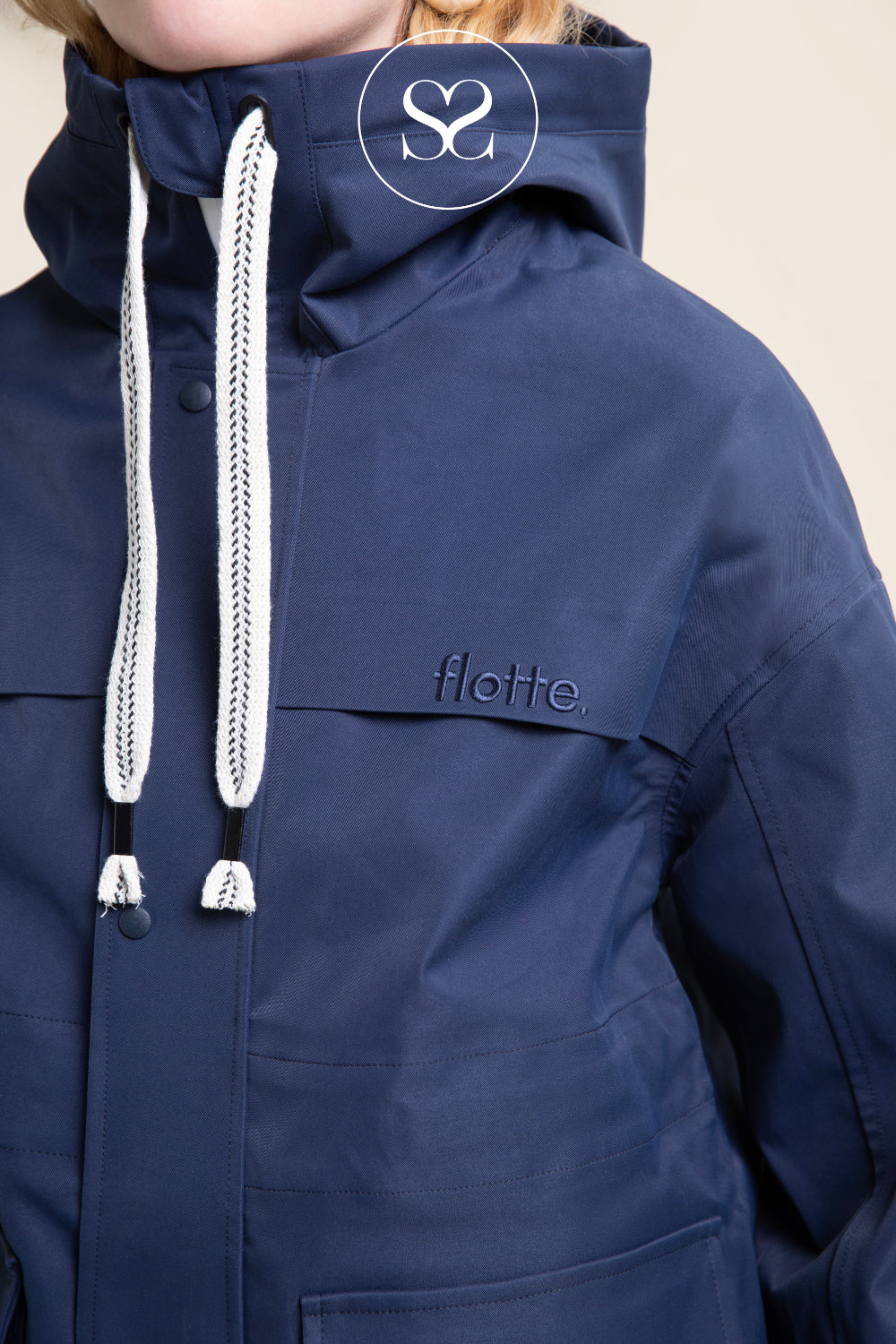 Close-up of a navy blue hooded funnel neck jacket with white braided drawstrings and a small tonal 'flotte.' logo on the chest, worn by a model against a light beige background.
