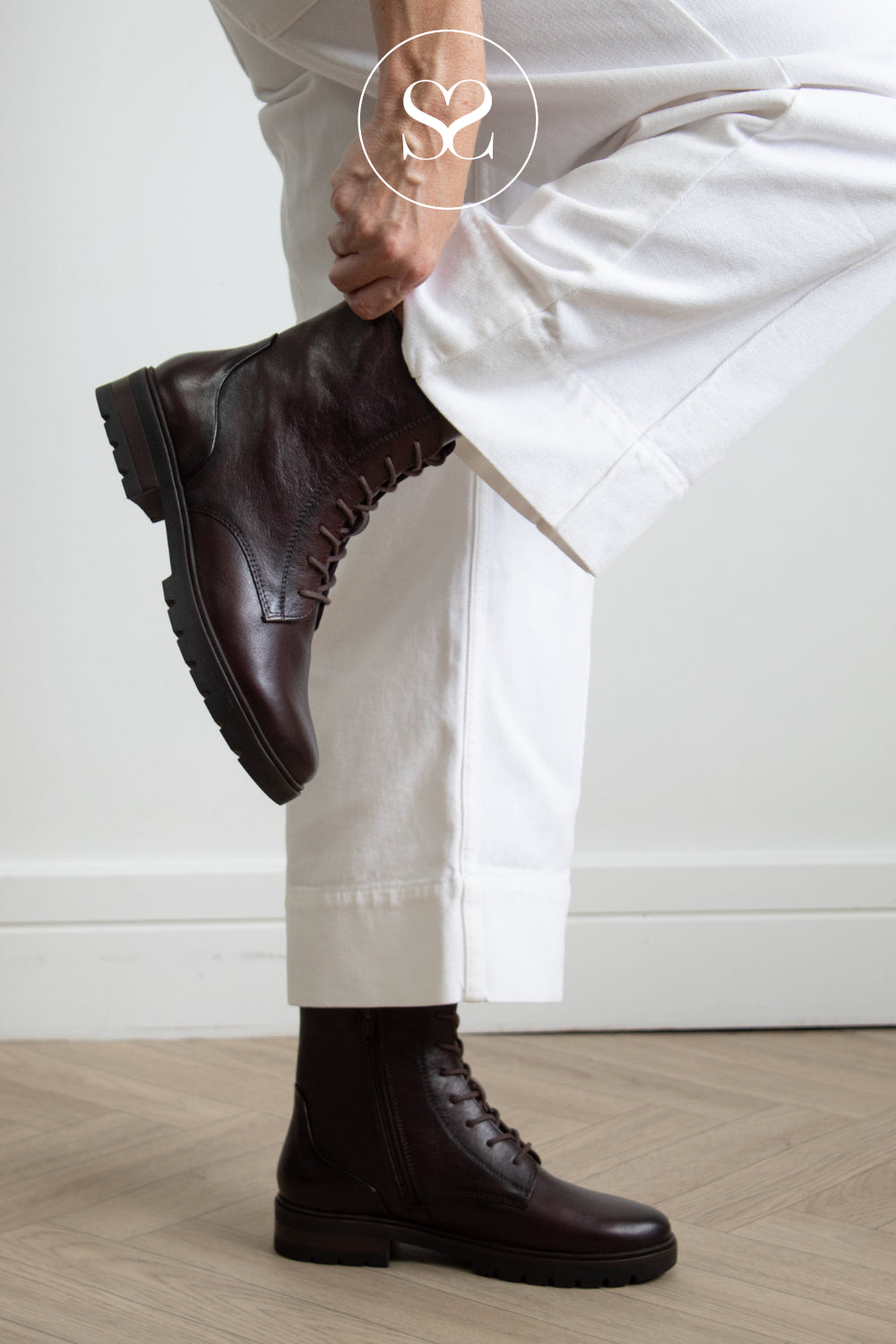 A person wearing Ara dark brown leather lace-up ankle boots and white trousers, lifting one boot to show the side profile against a clean white wall and light wood floor.