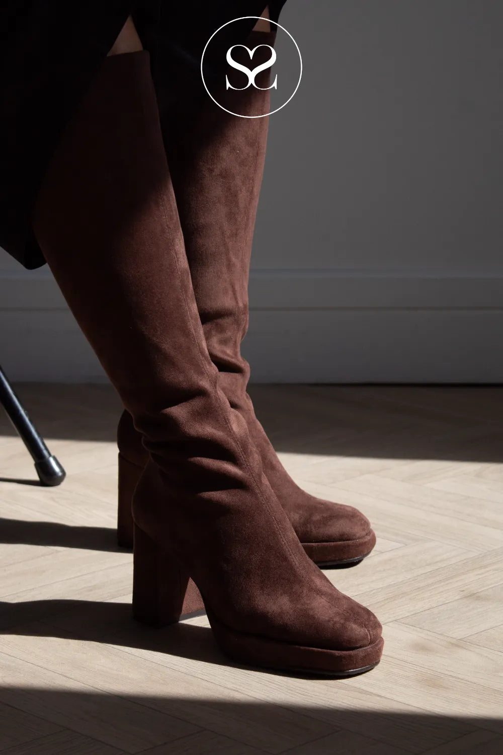 A pair of dark brown suede knee high boots with chunky block heels, shown on a model standing on a light wood herringbone floor in dramatic side lighting. A circular 'SS' logo is visible at the top.