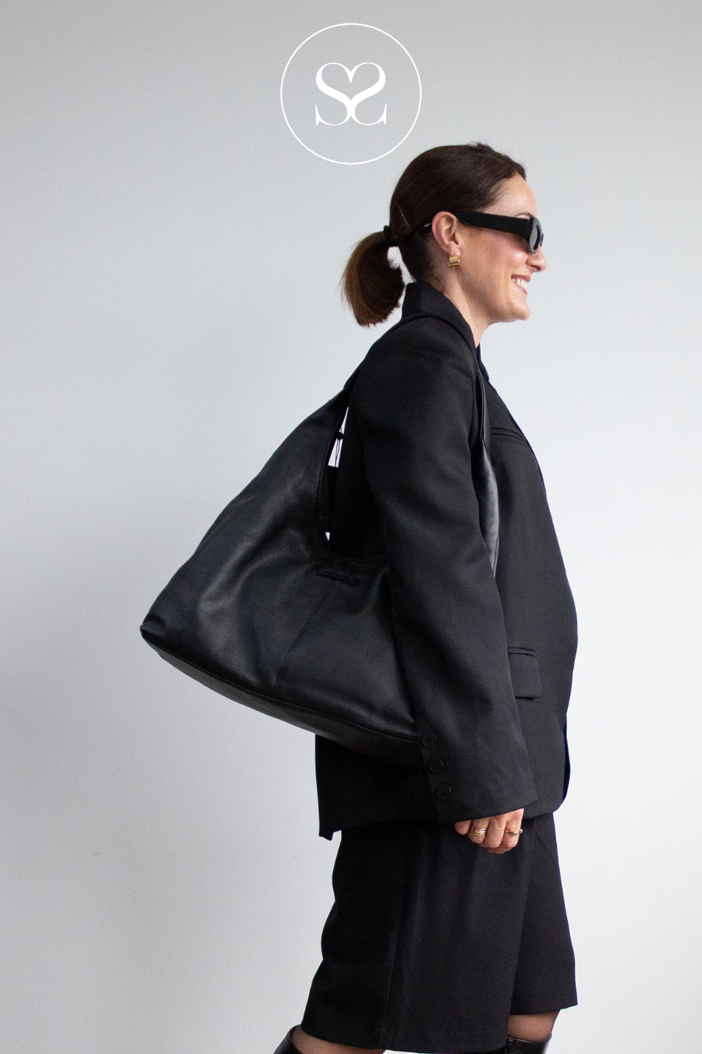 A woman in a black blazer, black skirt, and black sunglasses carries a large Depeche black leather shoulder bag against a light gray studio background. A circular logo for Sheneil Shoes is visible at the top center.