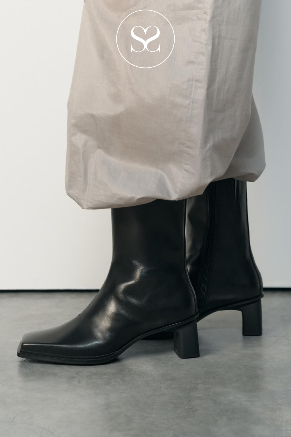 A pair of black leather ankle boots with a block heel and square toe from Vagabond, worn with loose light beige trousers. A white circular logo with an 'S' is printed on the trouser leg.
