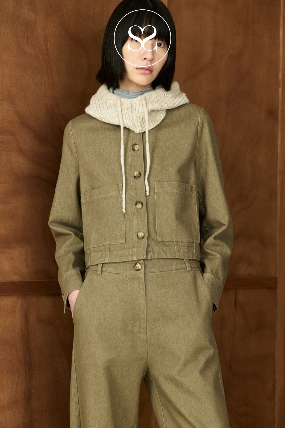 A model wearing a matching olive green cropped hooded jacket and wide-leg trousers set, styled with a cream knit neck warmer, against a warm wood-paneled background.