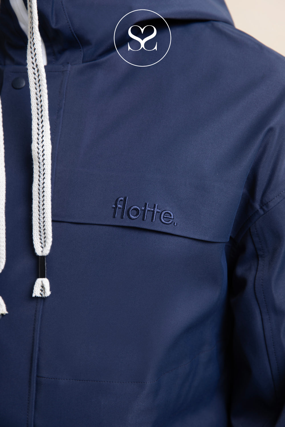 Close-up of a navy blue hooded jacket with white drawstrings and an embroidered 'flotte.' logo on the chest, shown against a light neutral background. froissart style