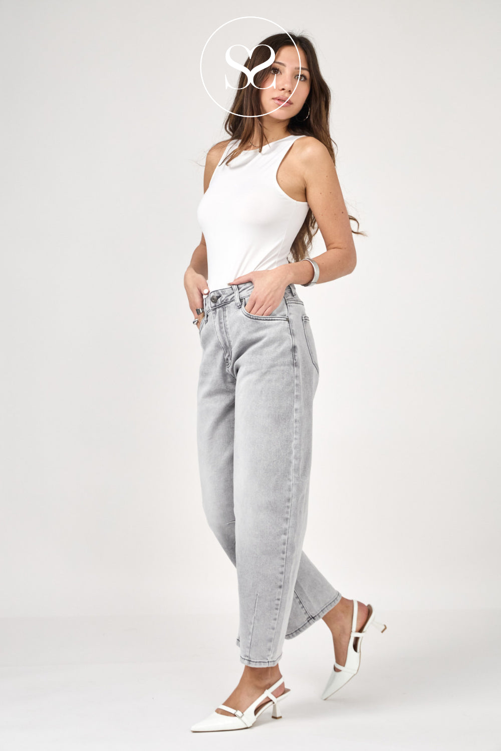 A woman modeling light-wash high-rise jeans, a white sleeveless top, and white pointed-toe slingback heels against a plain light gray studio background.