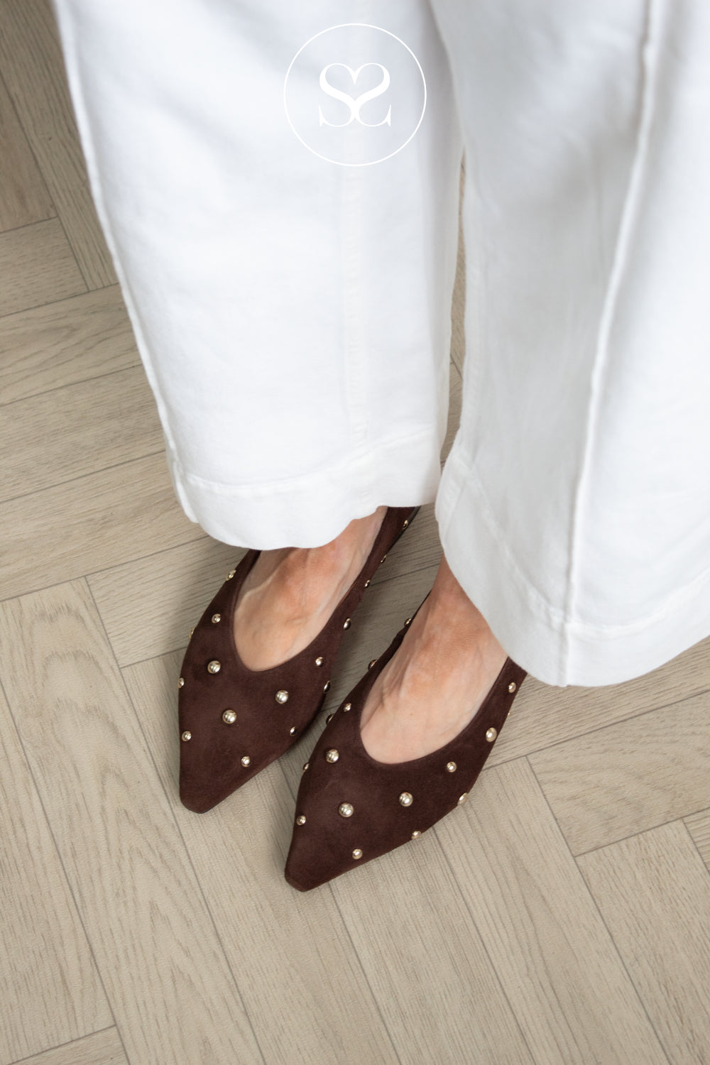 A close-up of a woman wearing angel alarcon dark brown suede pointed-toe flats with small gold studs, paired with white cropped pants, standing on a light wood herringbone floor.