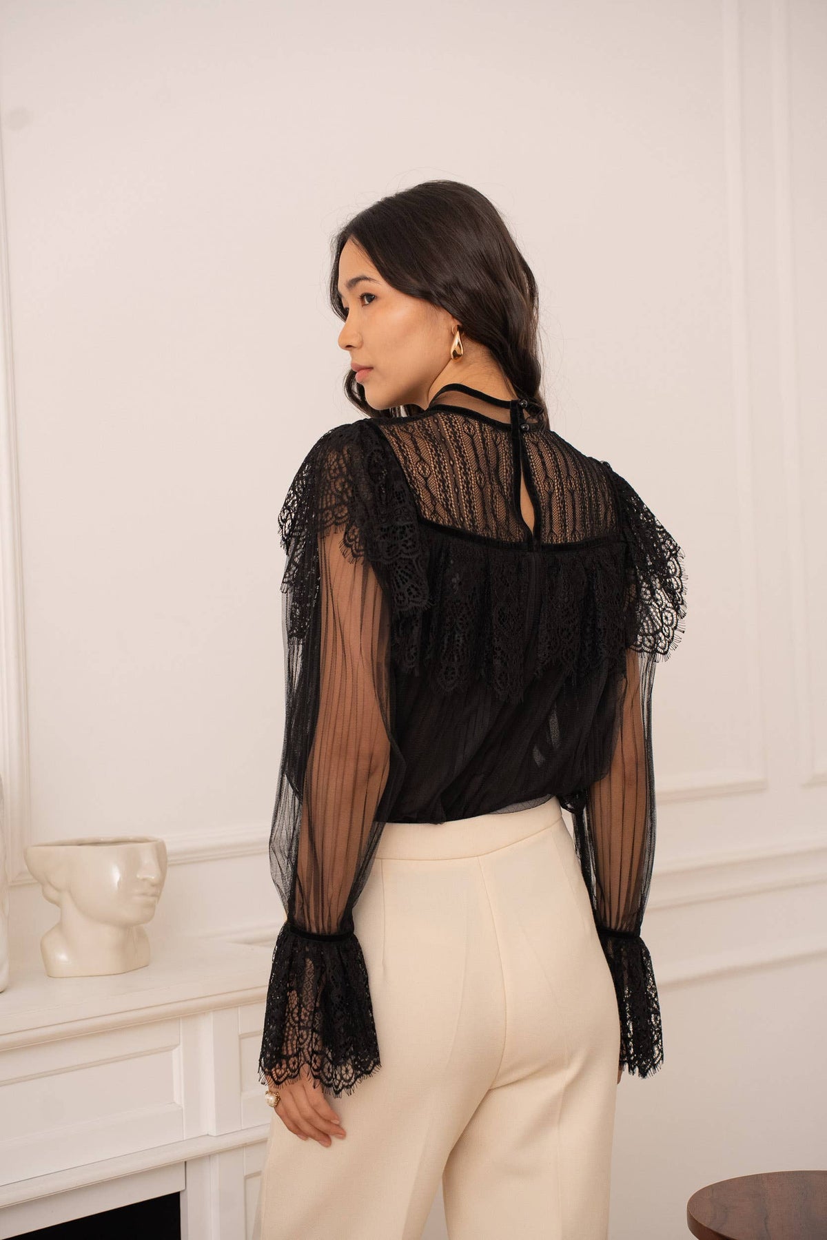 Woman wearing a black sheer blouse with lace details in a room with white walls.