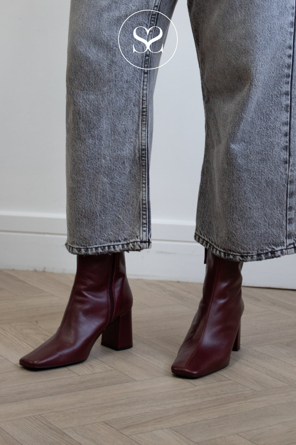 A woman wearing dark burgundy leather ankle boots with a block heel and square toe from Angel Alarcon, paired with faded gray jeans, standing on a light wood herringbone floor.