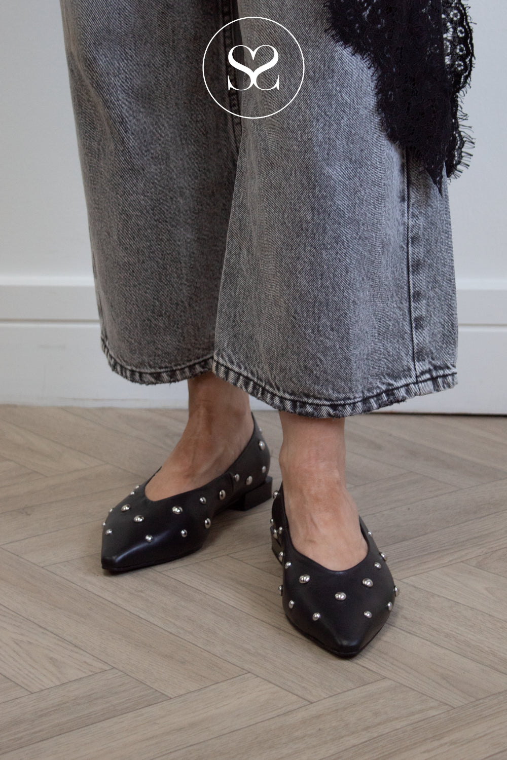 A person wearing black pointed-toe flats with silver studs from Angel Alarcon, paired with gray cropped jeans and a black lace top, standing on a light wood herringbone floor.