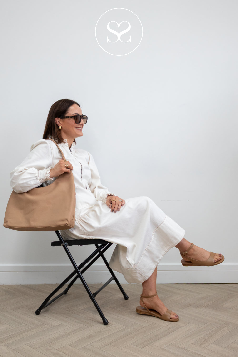 Woman in white outfit holding a beige tote bag, sitting on a black stool against a white wall wearing flat tan sandals from angel Alarcon