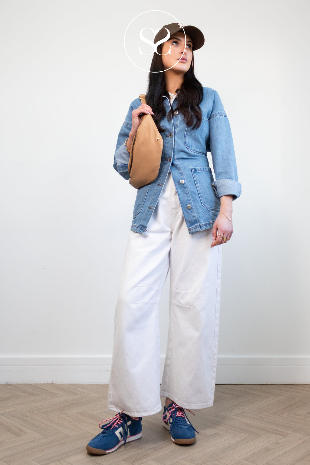 Person wearing a denim jacket, white pants, and blue sneakers from Back 70 , holding a beige bag.